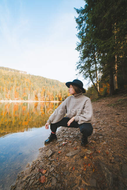 Joven turista femenina fresca en ropa de moda mirando lejos de la costa seca contra el agua transparente que refleja los árboles de otoño - foto de stock