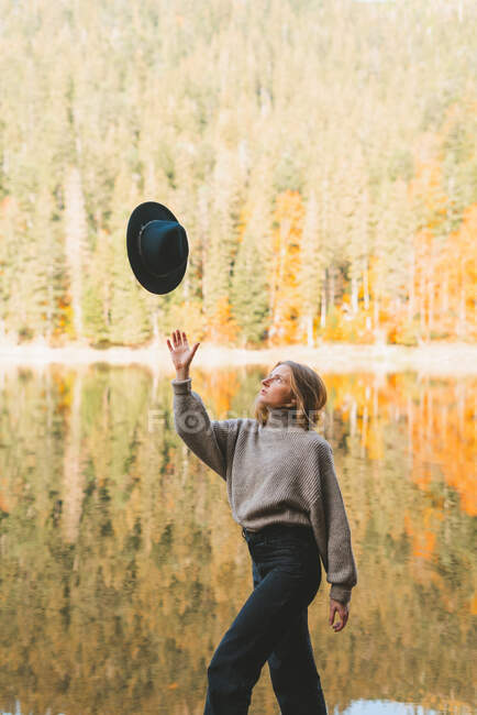 Young female traveler in casual outfit looking up at hat in air while strolling with raised arm against trees reflecting in water — Stock Photo