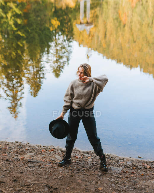 From above young blond cool female tourist in trendy apparel looking away while standing on coast against water reflecting trees — Stock Photo