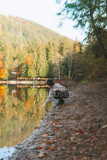 Side view of female traveler in casual apparel and hat reflecting in transparent water against trees in fall — Stock Photo