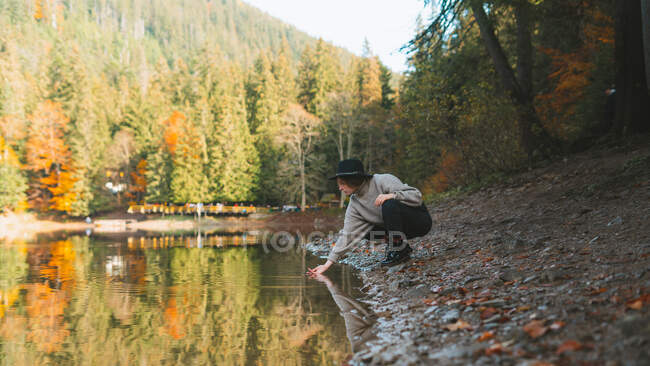 Seitenansicht einer Reisenden in lässiger Kleidung und Hut, die sich im transparenten Wasser gegen Bäume im Herbst spiegelt — Stockfoto