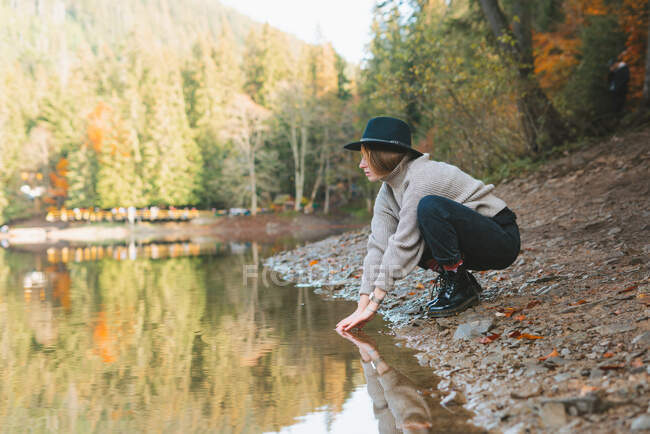 Side view of anonymous female traveler in casual apparel and hat reflecting in transparent water against trees in fall — Stock Photo