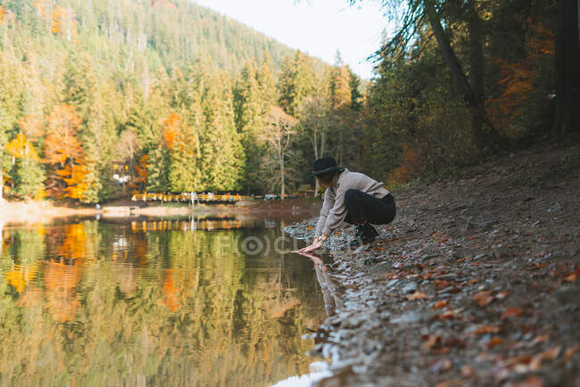 Side view of anonymous female traveler in casual apparel and hat reflecting in transparent water against trees in fall — Stock Photo