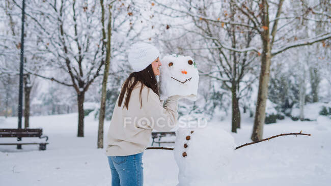 Vista lateral de una joven alegre hembra con muñeco de nieve en el parque con árboles sin hojas en invierno - foto de stock