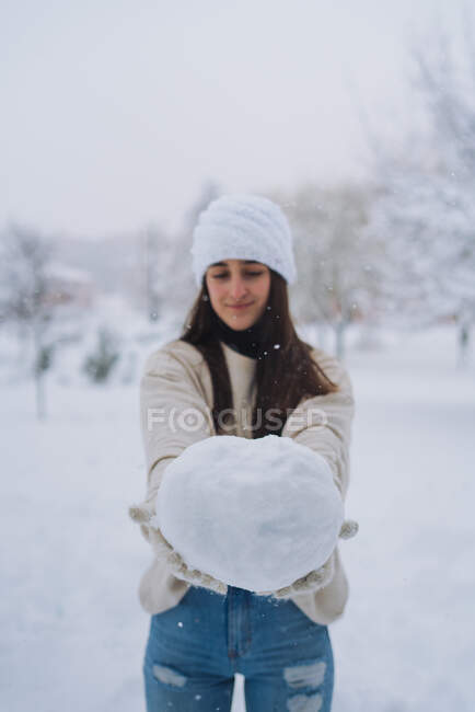 Young content ethnic female in knitted hat showing snowball under white sky in park of Madrid — Stock Photo