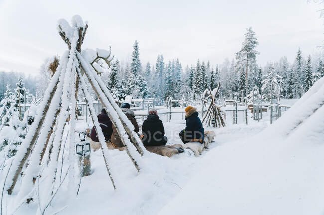 Вид сзади на неузнаваемых путешественников в теплой одежде и шляпах, сидящих у костра во время кемпинга в зимнем лесу, покрытом снегом — стоковое фото