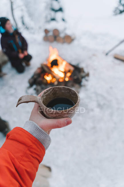 De cima de colheita anônimo viajante feminino em roupas quentes segurando xícara de barro de café quente perto de fogueira em terreno nevado no inverno — Fotografia de Stock