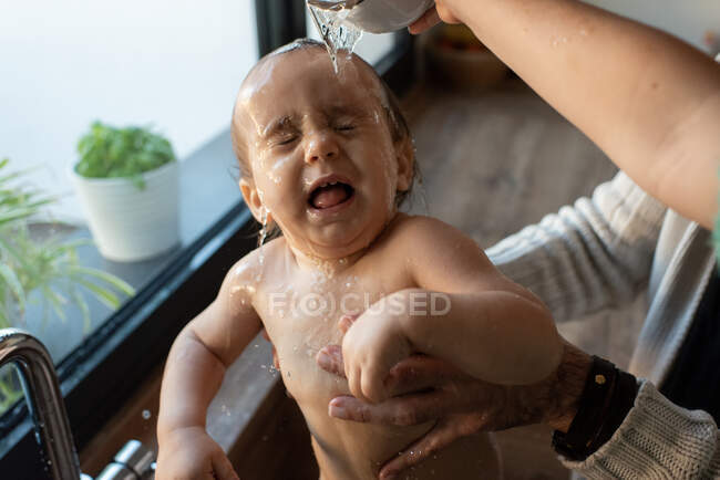 З висоти нерозпізнаної матері і батька купаються милий малюк в раковині на кухні, наливаючи воду на обличчя — стокове фото
