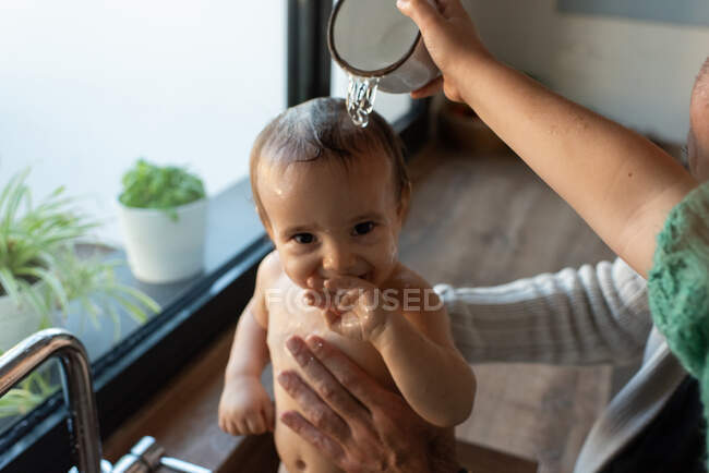 З понад врожаю анонімні мати і батько купаються милою усмішкою малюка в раковині на кухні і заливають водою по голові. — стокове фото