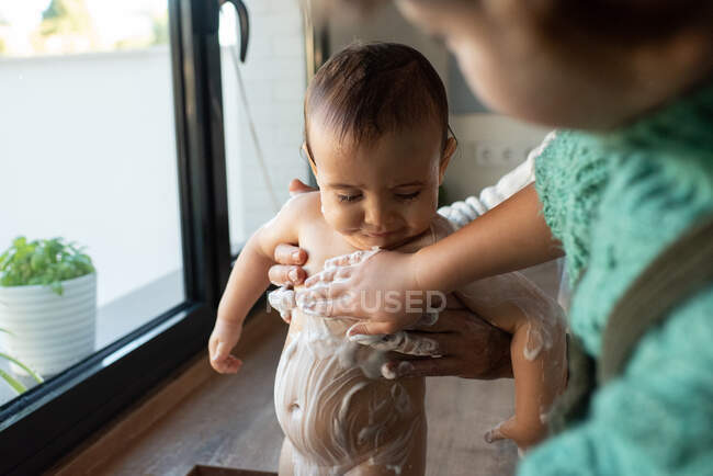 Delighted father and kids bathing cute baby full of foam in sink in kitchen while spending time together — Stock Photo