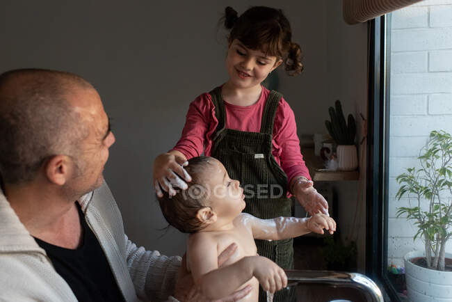 Маленькая девочка моет голову милого ребенка на руках отца во время купания в раковине на кухне — стоковое фото