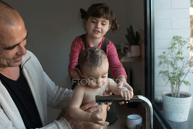 Little girl washing cute baby in arms of father during bathing in sink in kitchen — Stock Photo