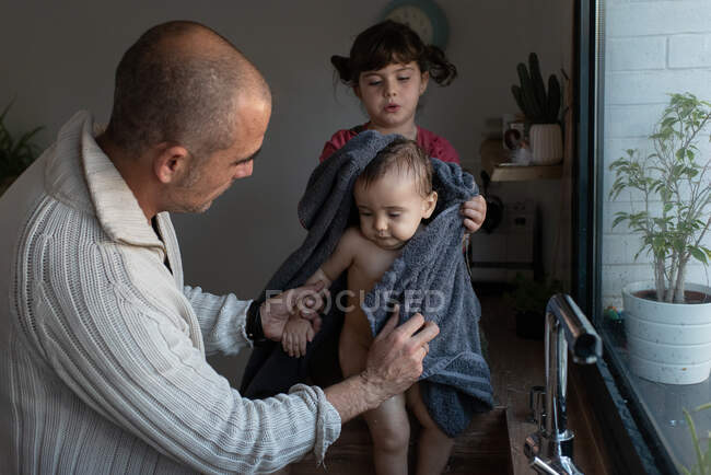 Padre y niña limpiando lindo niño desnudo con toalla después de bañarse en el fregadero en la cocina - foto de stock