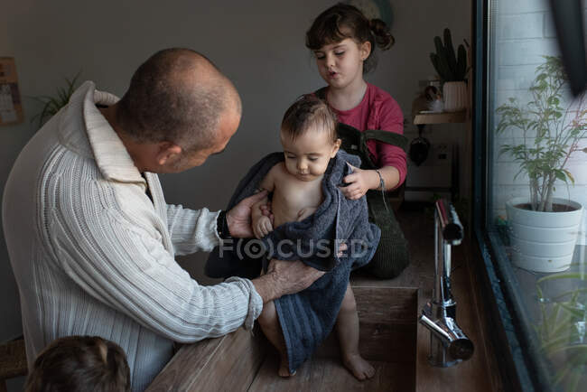 Батько і маленька дівчинка витирають милого голого малюка рушником після купання в раковині на кухні — стокове фото