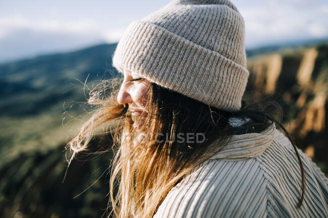 Vue latérale d'une jeune voyageuse joyeuse aux cheveux volants et aux yeux fermés contre des montures exposées au soleil — Photo de stock