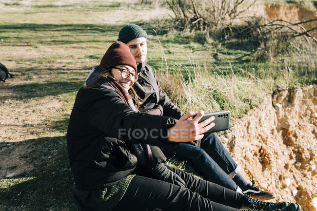 Seitenansicht eines glücklichen Touristenpaares in Oberbekleidung, das ein Selbstporträt auf dem Handy macht, während es sich auf einem Berg gegen eine Schlucht im Gegenlicht ausruht — Stockfoto