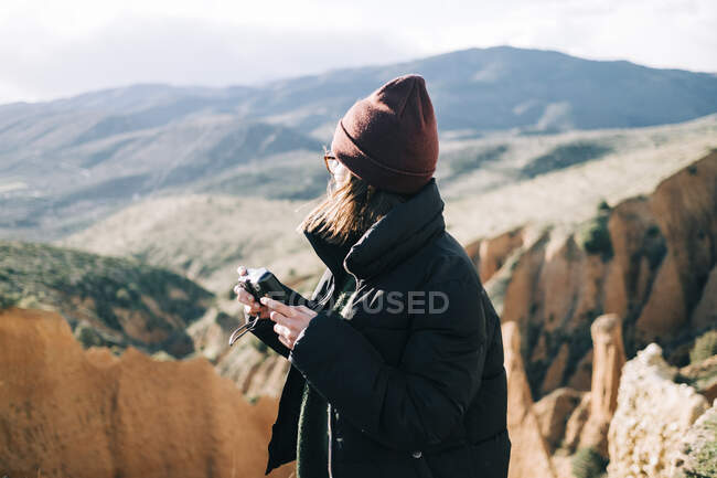 Seitenansicht einer unkenntlichen Touristin, die eine alte Kamera in der Hand hält, während sie im Sonnenlicht auf einer Schlucht vor einem Berg steht — Stockfoto
