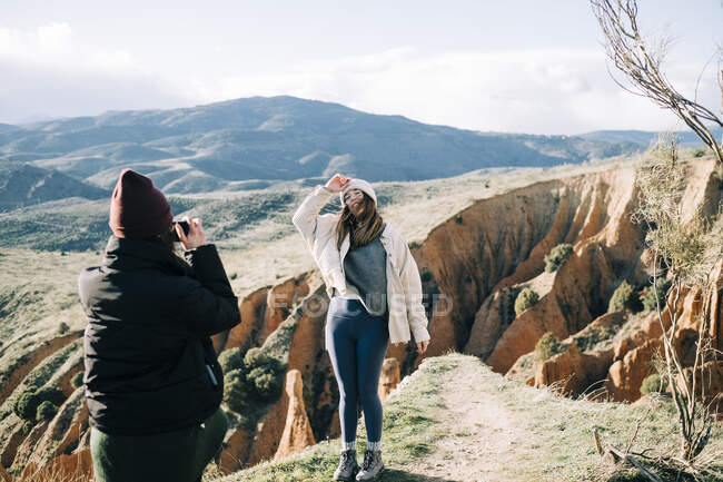 Невпізнавана жінка-туристка фотографує щасливого найкращого друга на камеру проти гори і каньйону на сонячному світлі — стокове фото