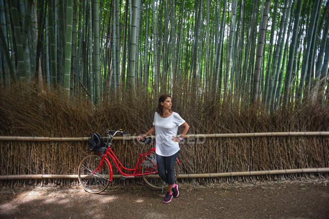 Attractive young caucasian woman with her bicycle looking up at trees in landmark Arashiyama Bamboo Grove forest in Kyoto, Japan — Stock Photo