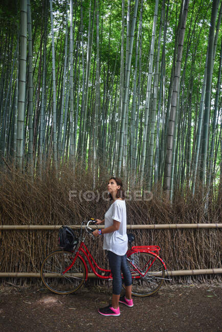 Attractive young caucasian woman with her bicycle looking up at trees in landmark Arashiyama Bamboo Grove forest in Kyoto, Japan — Stock Photo