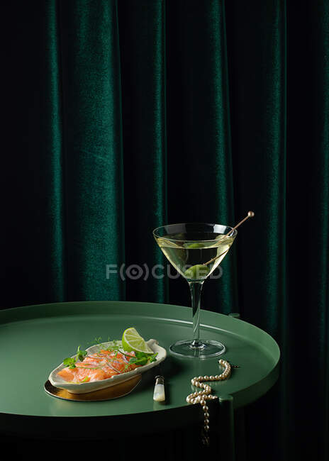 Сверху аппетитные ломтики копченого лосося подаются на тарелке с лаймом и луком и помещаются на круглый стол со стаканом вермута с оливками возле элегантного жемчужного ожерелья — стоковое фото