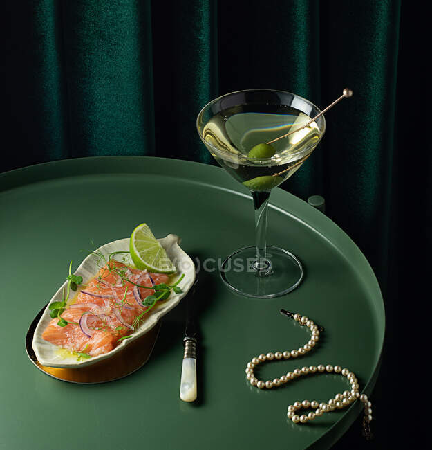 From above of appetizing smoked salmon slices served on plate with lime and onion and placed on round table with glass of vermouth with olive near elegant pearl necklace — Stock Photo