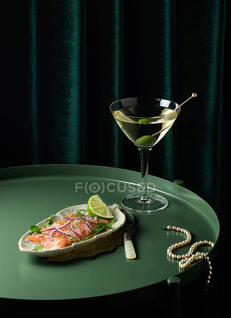 Зверху апетитні шматочки копченого лосося подаються на тарілці з лаймом і цибулею і кладуть на круглий стіл зі склянкою вермута з оливковою олією біля витонченого перлового намиста — стокове фото