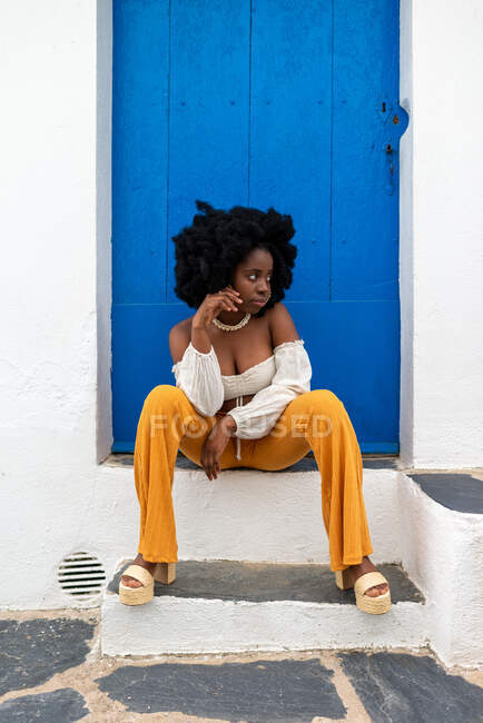 Giovane donna afroamericana in abito estivo alla moda seduto sulla soglia di casa vicino edificio sulla vecchia strada in città — Foto stock