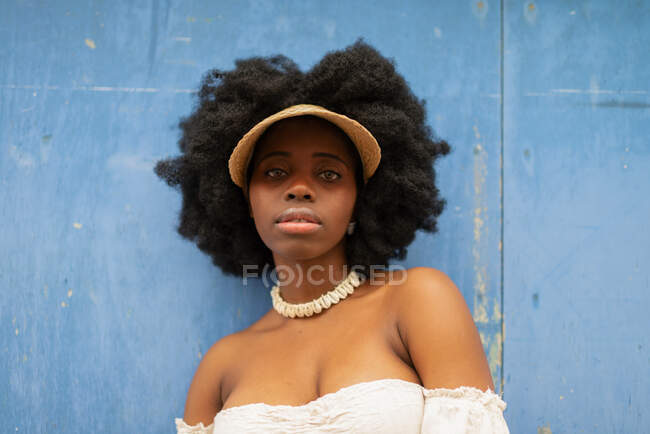 Low angle of carefree African American female with Afro hairstyle and in cap standing in street against shabby wall and looking at camera — Stock Photo