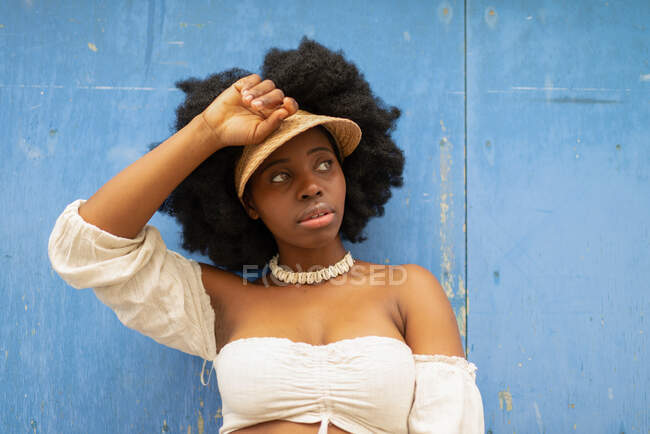Low angle of carefree African American female with Afro hairstyle and in cap standing in street against shabby wall and looking away — Stock Photo