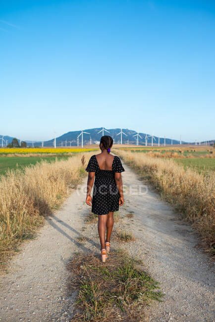 Rückansicht einer nicht wiedererkennbaren Afroamerikanerin im Sommerkleid, die ländliche Straße in der Landschaft vor dem Hintergrund von Sonnenblumenfeld und Bergen entlanggeht — Stockfoto
