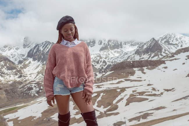African American female in stylish outfit standing on background of snowy mountains in winter — Stock Photo