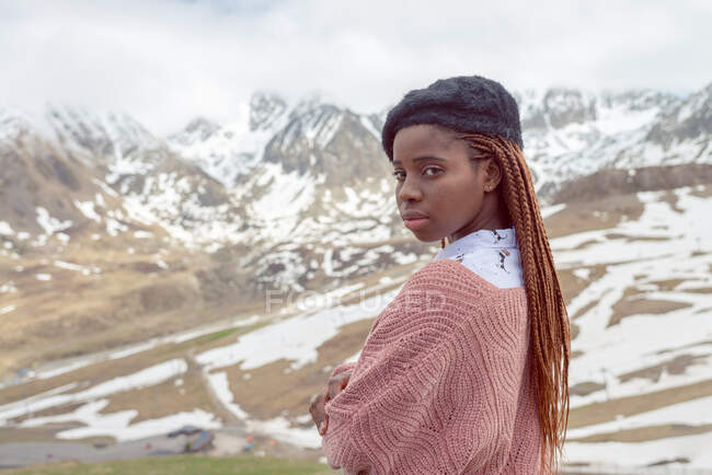 Seitenansicht einer Afroamerikanerin in stylischem Outfit, die im Winter vor dem Hintergrund verschneiter Berge steht — Stockfoto