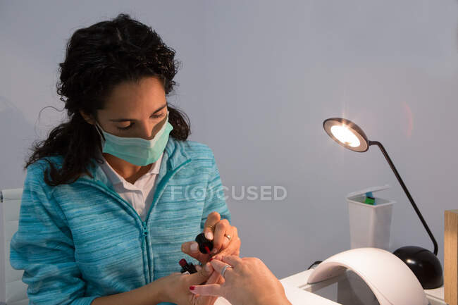 Manicure feminino atento na máscara facial aplicando laca no prego da mulher de colheita perto da lâmpada brilhante no salão de beleza — Fotografia de Stock