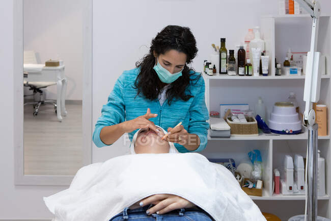 Mestre de beleza irreconhecível aplicando cera quente com espátula no rosto de mulher deitada durante o processo de depilação no salão de spa — Fotografia de Stock