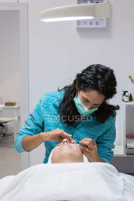 Анонимный косметолог в маске, лечит взрослую женщину с закрытыми глазами во время процедуры по уходу за лицом в салоне красоты — стоковое фото