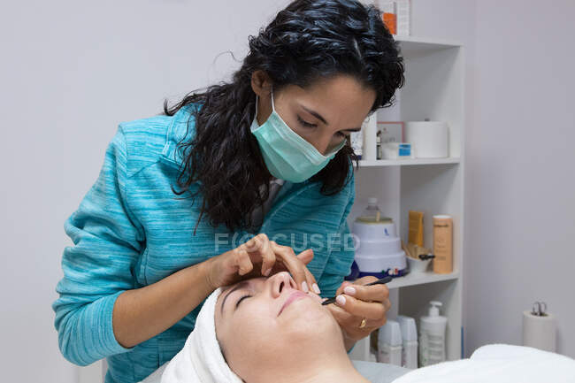 Cosecha esteticista anónima en la máscara de tratar a la mujer adulta con los ojos cerrados durante el procedimiento facial en el centro de belleza - foto de stock