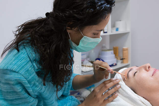 Cosmetólogo anónimo de cultivo aplicando cera caliente con aplicador en la ceja de la mujer con los ojos cerrados durante el procedimiento de depilación en el centro de spa - foto de stock