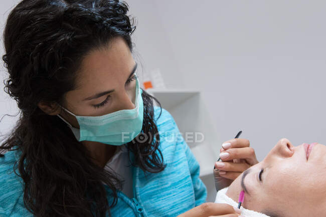Crop anonymous beautician in mask combing eyebrow of adult woman with closed eyes using brush during facial procedure in beauty center — Stock Photo