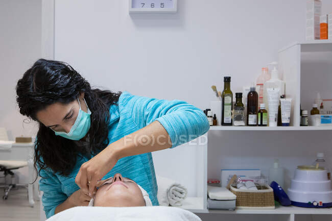 Schnitte anonyme Kosmetikerin in Maske Behandlung erwachsener Frau mit geschlossenen Augen während Gesichtsbehandlung im Schönheitszentrum — Stockfoto