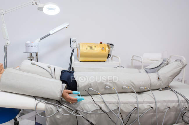 Seitenansicht einer anonymen Frau, die während der Pressotherapie im Schönheitssalon unter einer glänzenden Lampe auf dem Bett liegt — Stockfoto