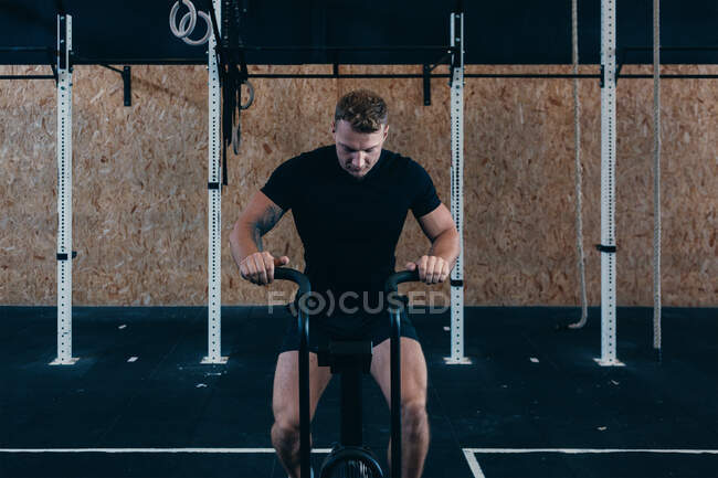 Atleta masculino serio sentado en bicicleta de aire mientras entrena en el gimnasio moderno durante el entrenamiento funcional - foto de stock