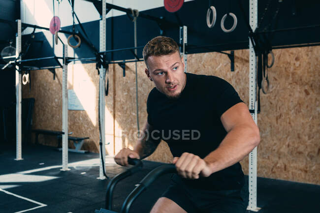 Serious male athlete sitting on air bike while training in modern gym during functional workout — Stock Photo