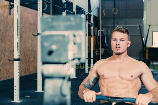 Focused male athlete with strong naked torso sitting on rowing machine and doing exercises during functional workout — Stock Photo