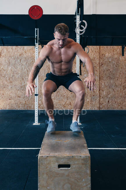 Muscular male athlete with naked torso jumping on wooden box during intense functional workout in fitness center — Stock Photo