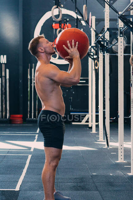 Vista lateral del deportista con torso desnudo lanzando balón medicinal durante el entrenamiento funcional en el gimnasio - foto de stock