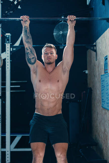 Serious shirtless male athlete hanging on horizontal bar while doing pull ups during functional workout in fitness center and looking at camera — Stock Photo
