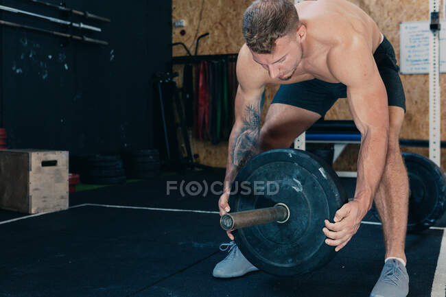 Deportista muscular con cuerpo desnudo fuerte poniendo placa de peso pesado en la barra mientras se prepara para el entrenamiento funcional en el gimnasio - foto de stock