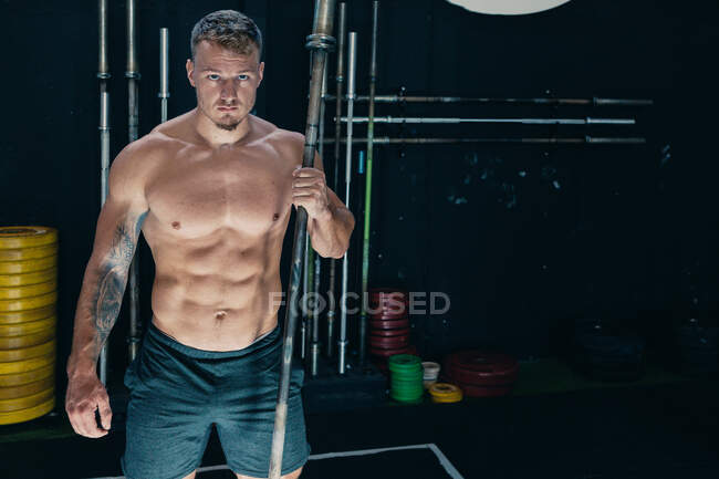 Homme déterminé avec corps nu athlétique debout avec haltère en métal dans la salle de gym sombre et regardant la caméra — Photo de stock