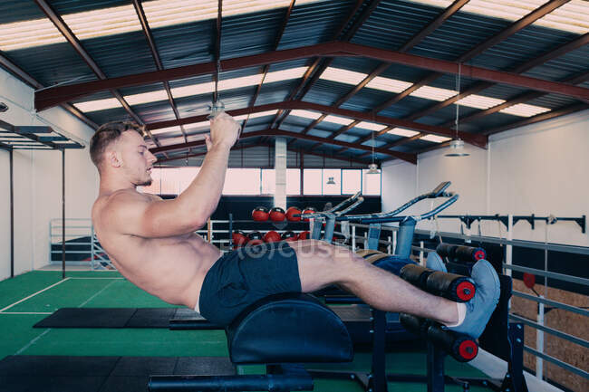 Vue latérale d'un athlète masculin déterminé avec torse nu faisant des craquements abdominaux sur banc pendant l'entraînement fonctionnel dans la salle de gym — Photo de stock